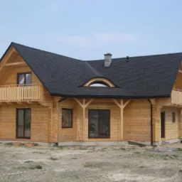 Nowoczesny dom z bali z czarnym dachem dwuspadowym, drewnianymi balkonami i nietypowym oknem w kształcie półksiężyca nad wejściem, otoczony niezagospodarowanym terenem.