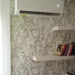 Biały klimatyzator ścienny z wyświetlaczem temperatury ustawiony na 22 stopnie Celsjusza, zamontowany na ścianie z tapetą w motywy drzew, obok półki z książkami.