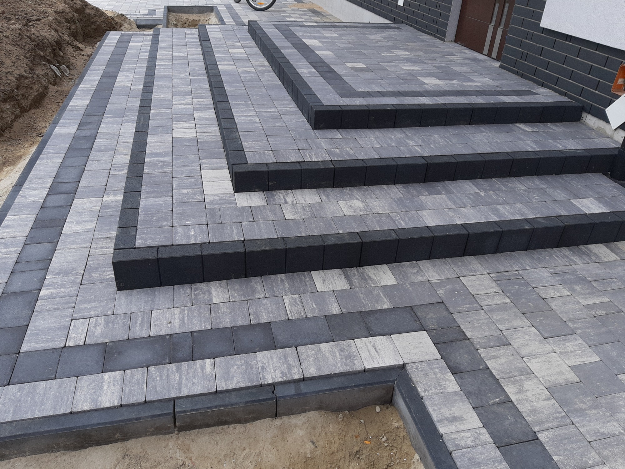 Ułożona kostka brukowa w odcieniach szarości i czerni tworząca schody wejściowe do budynku, z widocznymi elementami niedokończonej podsypki.