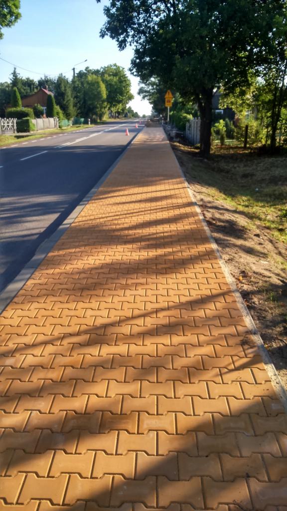 Nowy, pomarańczowy chodnik z kostki brukowej w kształcie litery H, biegnący wzdłuż drogi, z widocznymi drzewami i domami w tle, oświetlony jasnym słońcem.