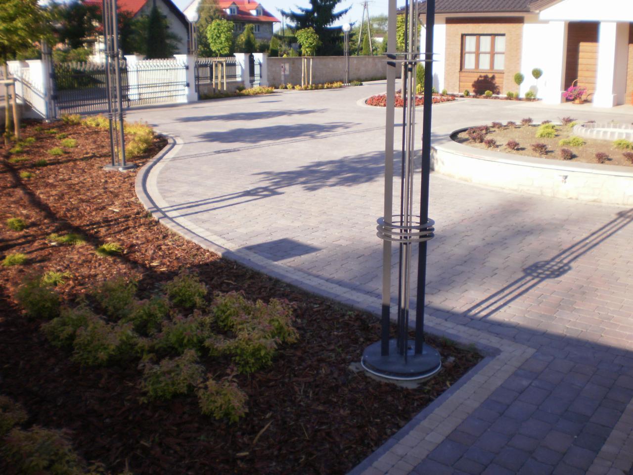 Ułożona kostka brukowa w odcieniach szarości tworzy elegancki podjazd. Widoczne rabaty z korą i niskimi krzewami oraz nowoczesne lampy.