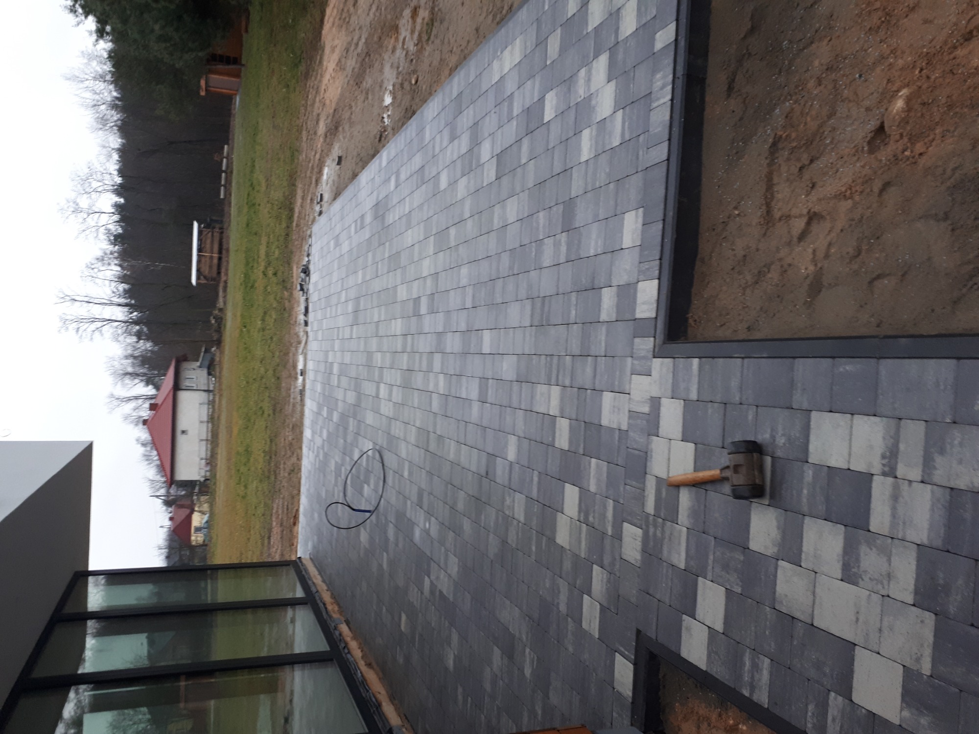 Ułożona szara kostka brukowa z gumowym młotkiem leżącym na powierzchni, widok z góry, tło z zielenią i budynkami.