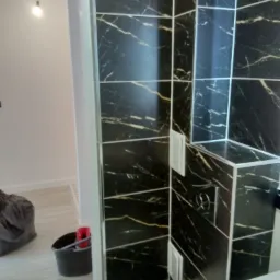 Ciemne, marmurowe płytki w łazience z widocznym fragmentem toalety i umywalki, w tle wiadro i narzędzia sugerujące prace wykończeniowe.