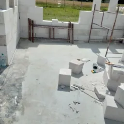 Wznoszenie ścian z bloczków betonu komórkowego; widoczne fragmenty rusztowania, narzędzia murarskie i butelka wody mineralnej na betonowej posadzce.