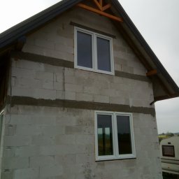 STAN -BUD - Surowy stan domu z białych bloczków, widoczne okna z białymi ramami i fragment dachu z drewnianą konstrukcją. W tle przyczepa kempingowa i zieleń.