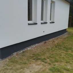 STAN -BUD - Boczny widok domu z trzema oknami i pomalowaną na szaro podmurówką. Świeżo otynkowana, biała elewacja. Widoczne fragmenty trawnika i drzew.