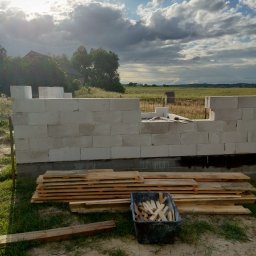 STAN -BUD - Ściana z betonu komórkowego w trakcie budowy domu, widoczne deski i pojemnik z drewnianymi elementami. W tle krajobraz z budynkiem i zachmurzone niebo.