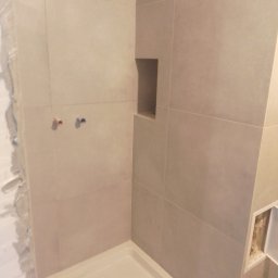 STAN -BUD - Wykończona wnęka w kabinie prysznicowej z beżowymi, dużymi płytkami. Widoczne wystające rury. Fragment ściany z niedokończonym wykończeniem.