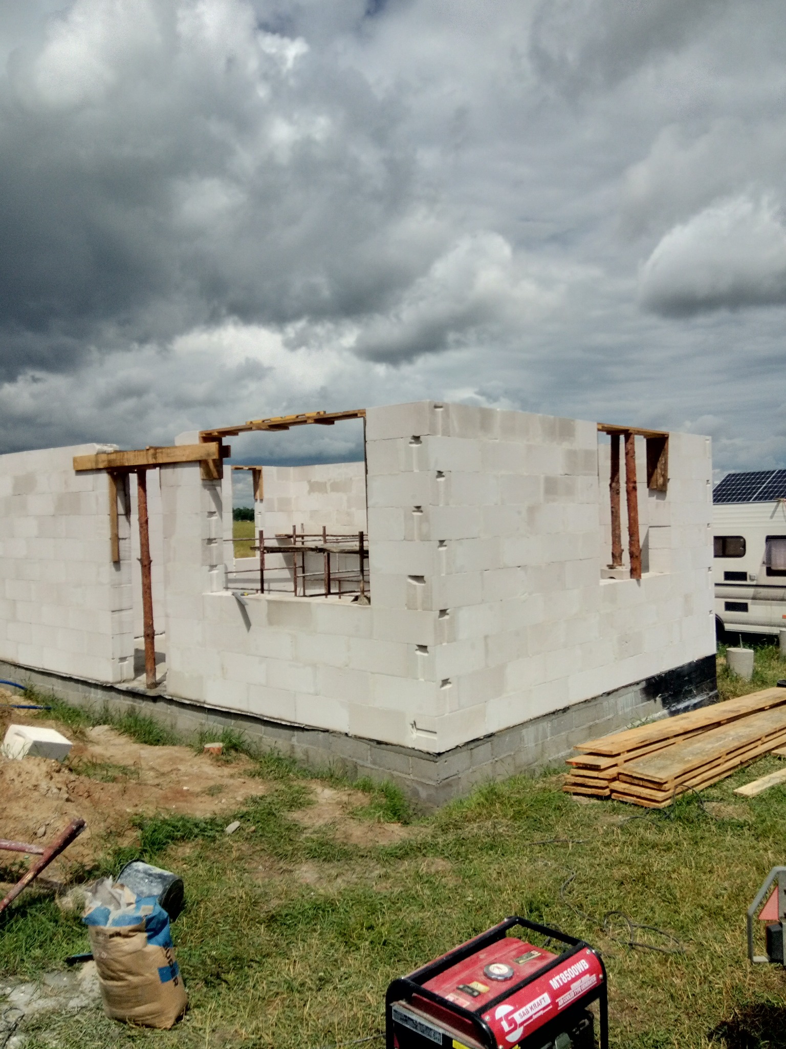 Wznoszenie ścian domu z betonu komórkowego, widoczne drewniane stemple podtrzymujące nadproża okienne, generator prądu na trawie.