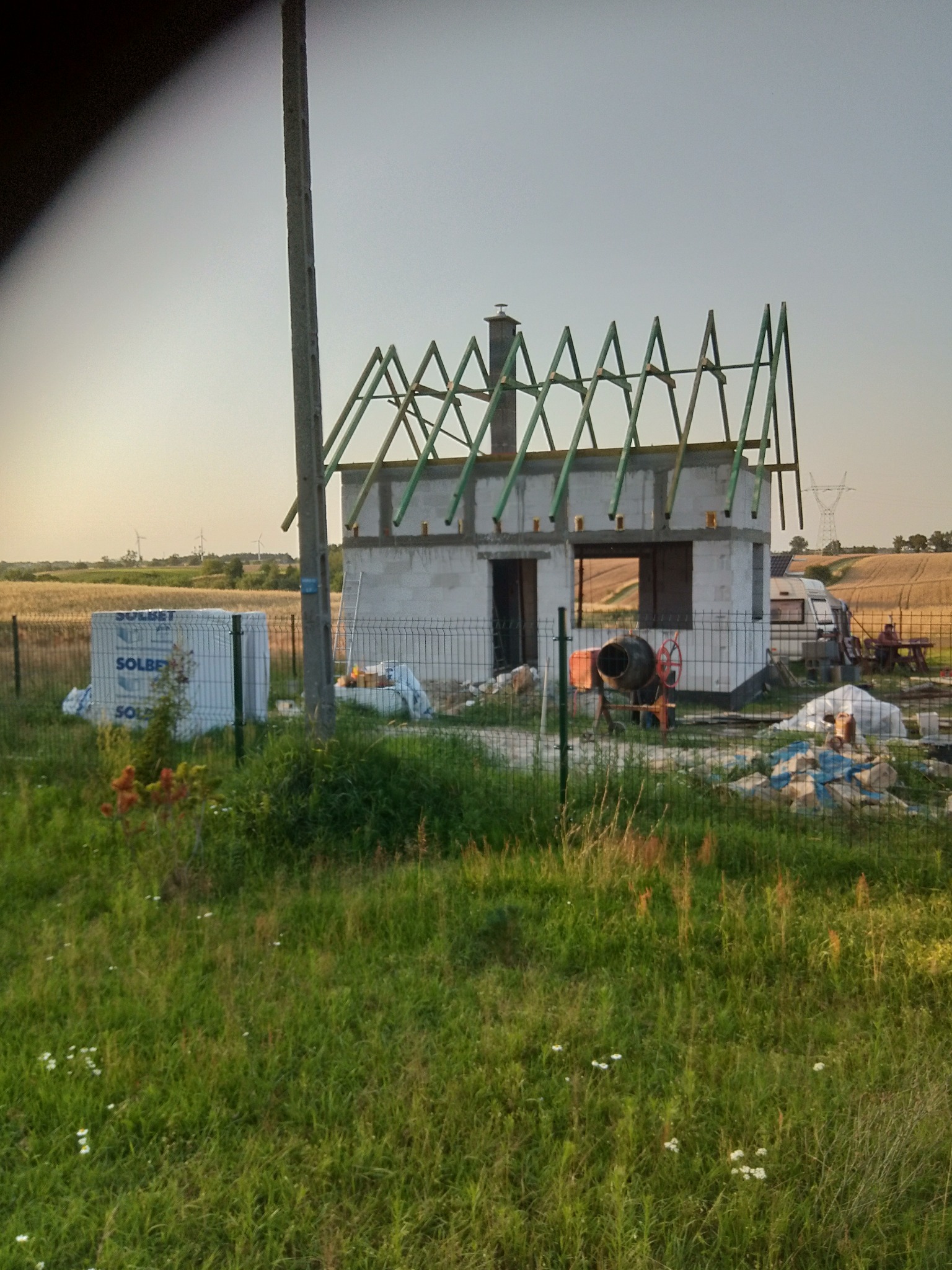 Surowy stan domu w budowie z widocznymi bloczkami, częściowo zadaszony konstrukcją więźby dachowej, na tle pola i błękitnego nieba.