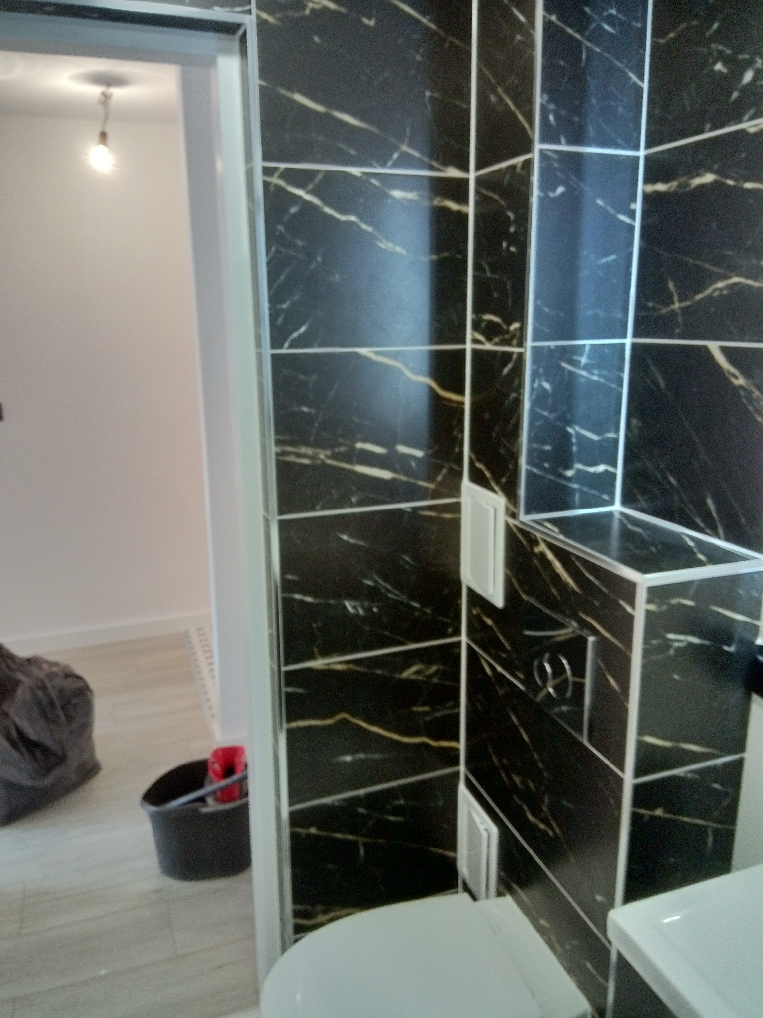 Ciemne, marmurowe płytki w łazience z widocznym fragmentem toalety i umywalki, w tle wiadro i narzędzia sugerujące prace wykończeniowe.