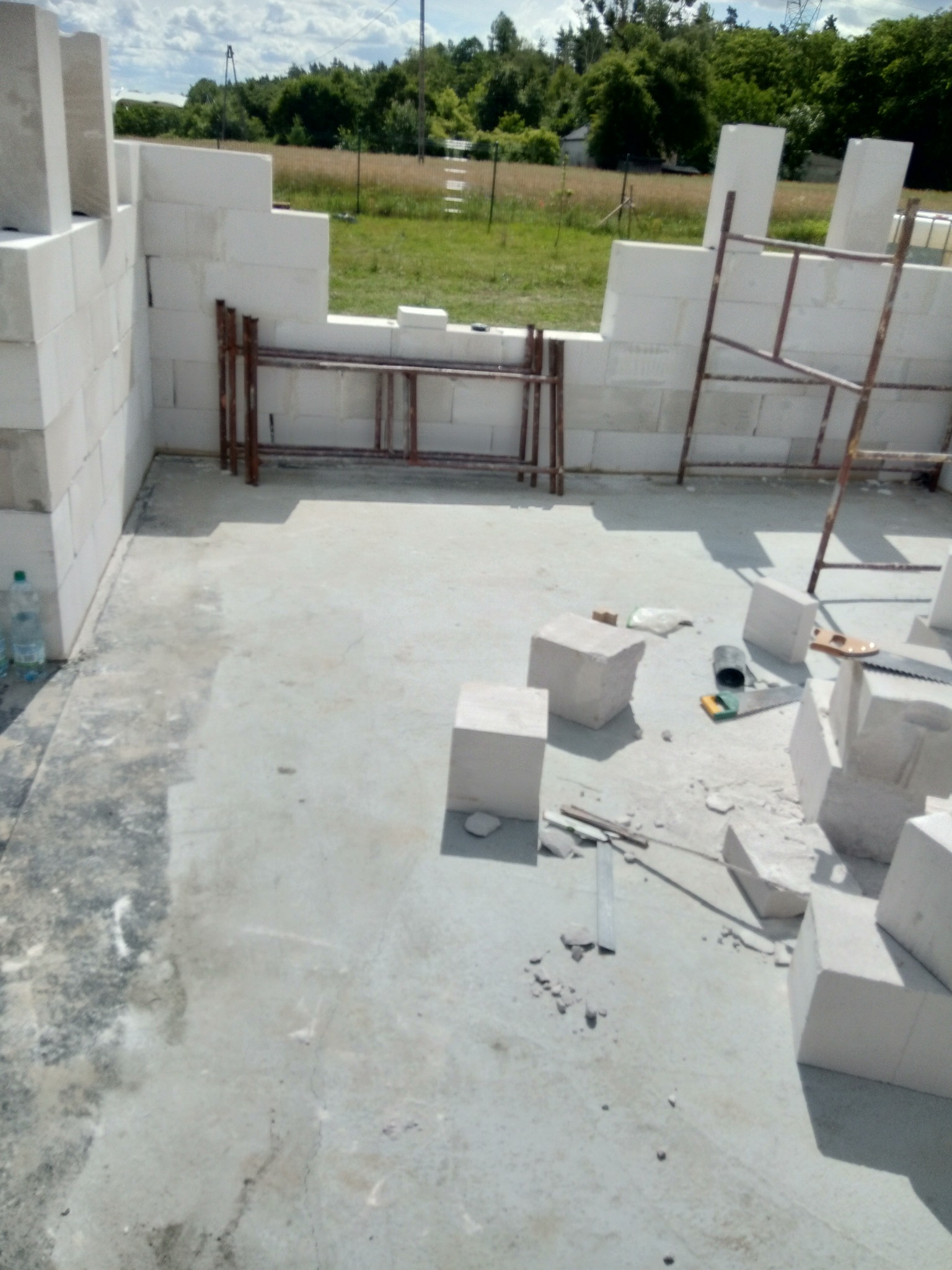 Wznoszenie ścian z bloczków betonu komórkowego; widoczne fragmenty rusztowania, narzędzia murarskie i butelka wody mineralnej na betonowej posadzce.