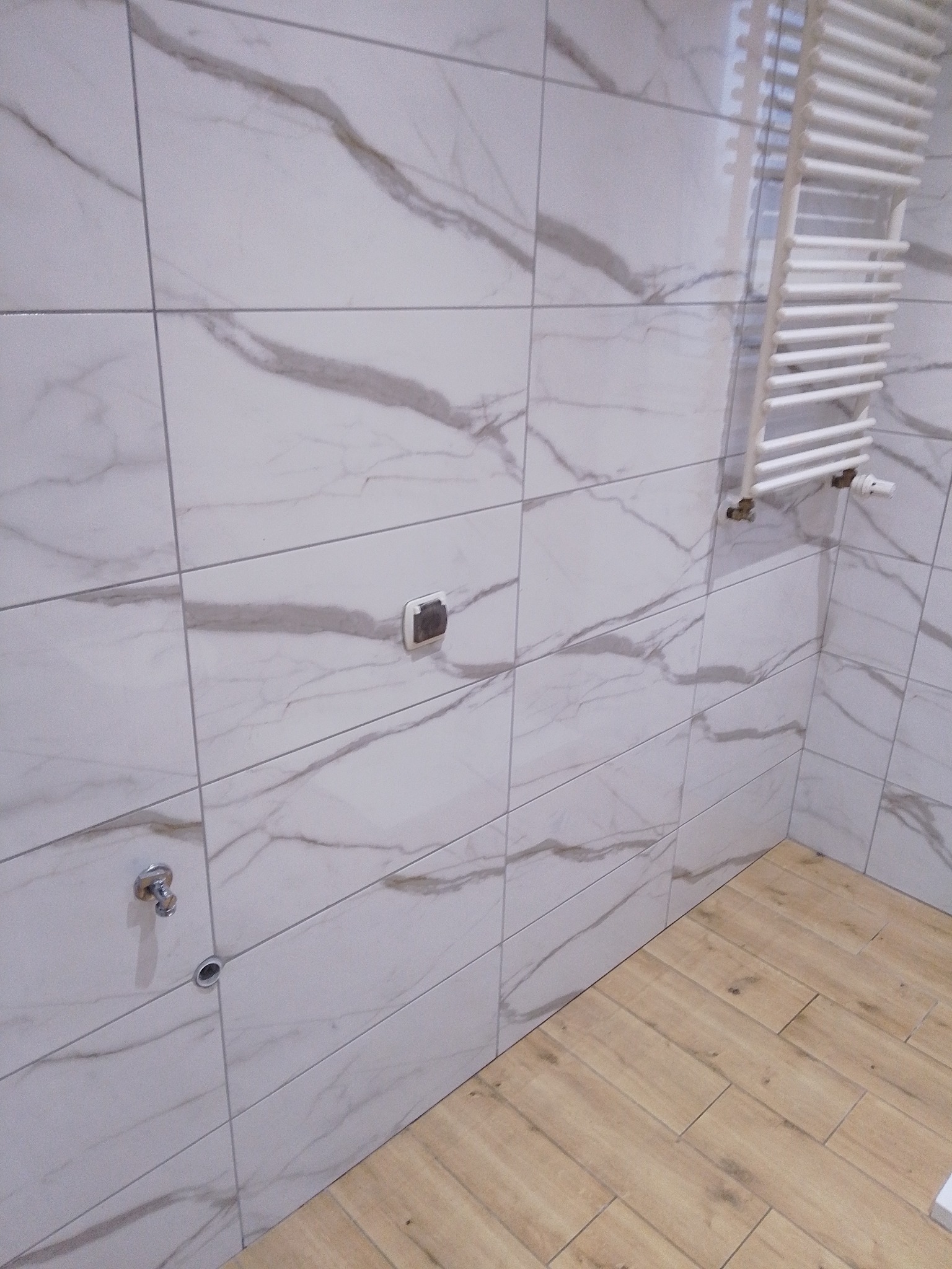 Wykończona łazienka z białymi płytkami imitującymi marmur i drewnopodobną podłogą. Widoczny grzejnik drabinkowy i elementy instalacji wodnej.