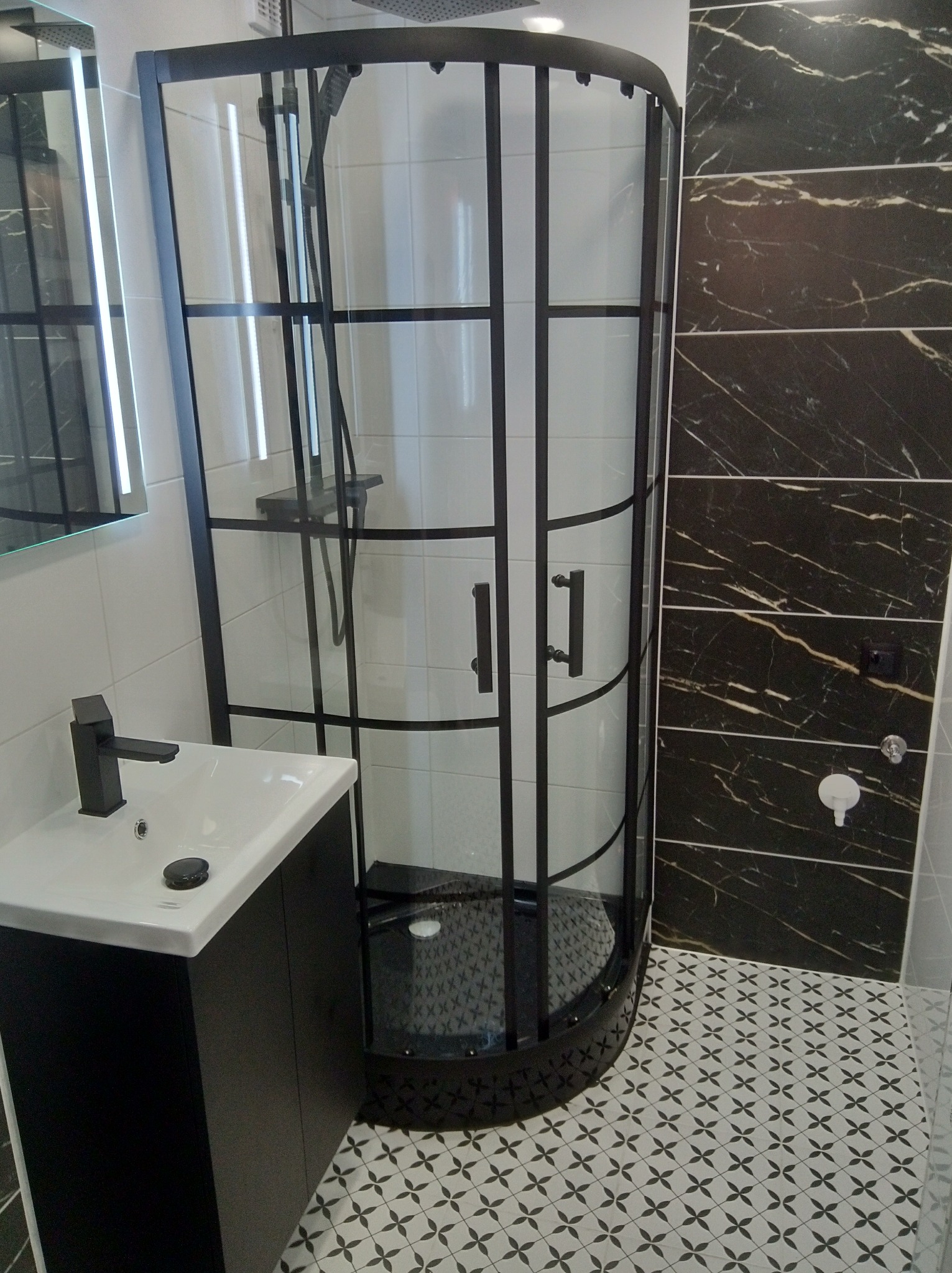 Nowoczesna kabina prysznicowa z czarną ramą i armaturą, umywalka z szafką w czerni, marmurowe płytki na ścianie i wzorzysta podłoga w wykończonej łazience.
