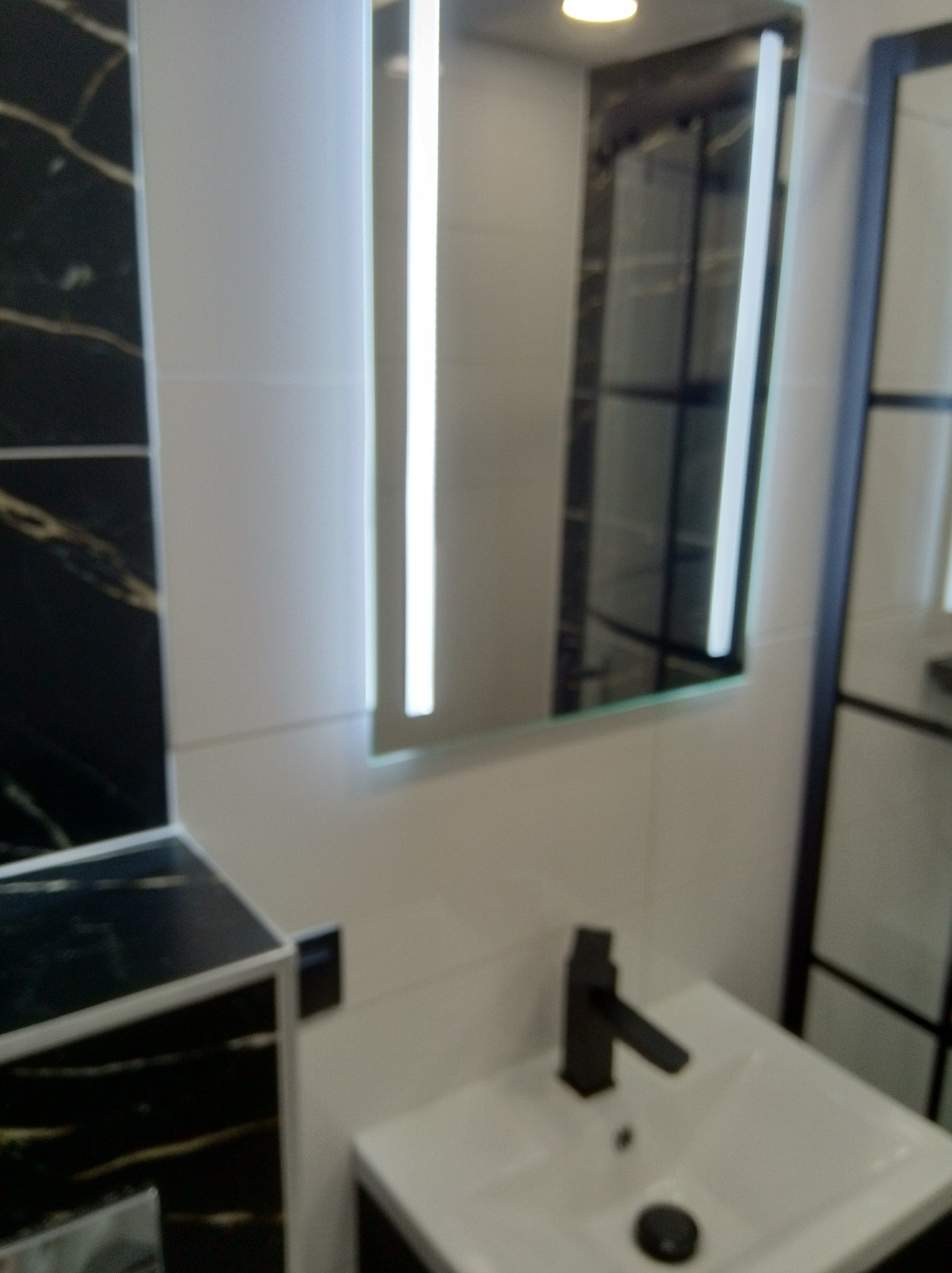 Nowoczesna łazienka z podświetlanym lustrem, czarną armaturą i marmurowymi akcentami. Minimalistyczny design, białe płytki i eleganckie wykończenie.