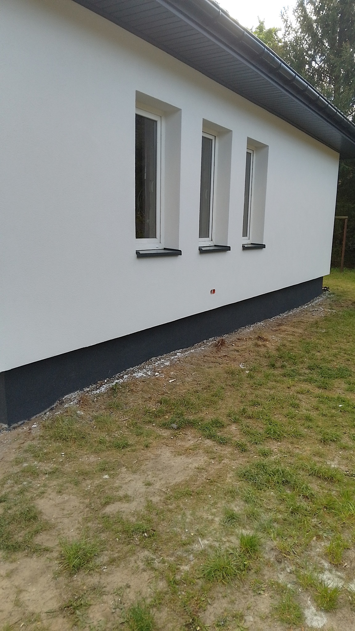 Boczny widok domu z trzema oknami i pomalowaną na szaro podmurówką. Świeżo otynkowana, biała elewacja. Widoczne fragmenty trawnika i drzew.