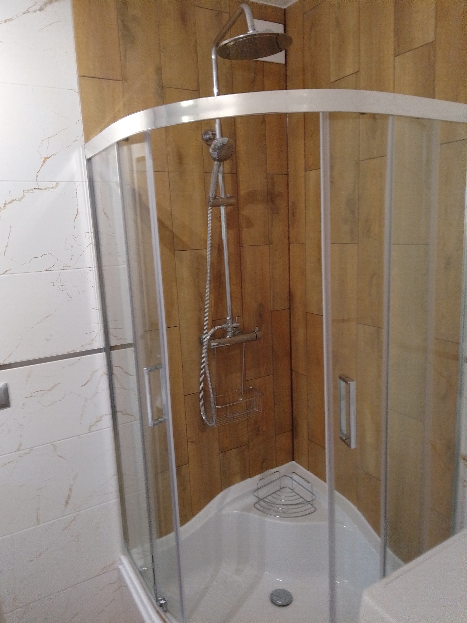 Narożna kabina prysznicowa z chromowanymi detalami i deszczownicą, wykończona płytkami imitującymi drewno i marmur. Widoczny odpływ i półka narożna.