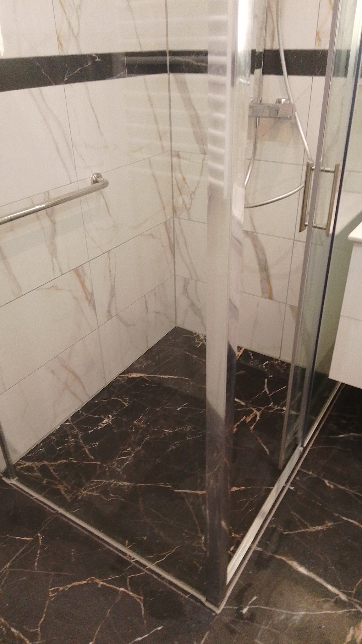 Nowoczesna kabina prysznicowa z marmurową posadzką i panelami ściennymi w odcieniach bieli i brązu. Chromowana armatura i szklane drzwi.