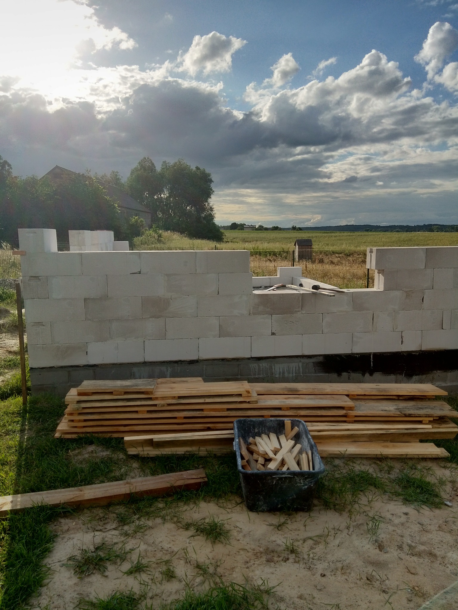 Ściana z betonu komórkowego w trakcie budowy domu, widoczne deski i pojemnik z drewnianymi elementami. W tle krajobraz z budynkiem i zachmurzone niebo.