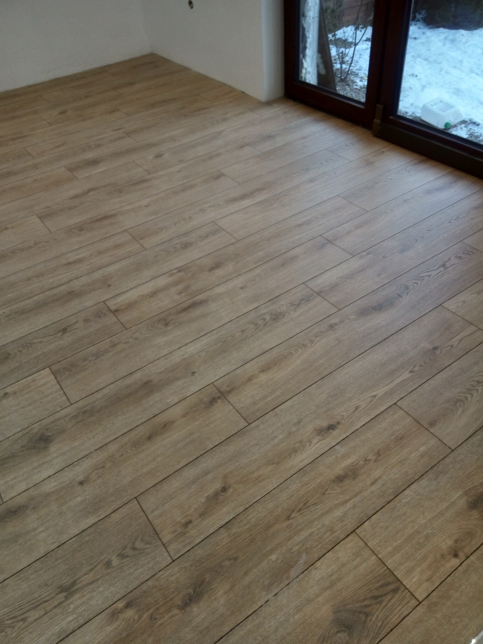 Ukończona podłoga z paneli w odcieniach brązu, ułożona wzdłuż okna balkonowego z widokiem na zimowy krajobraz. Widoczne łączenia paneli i fragment ściany.