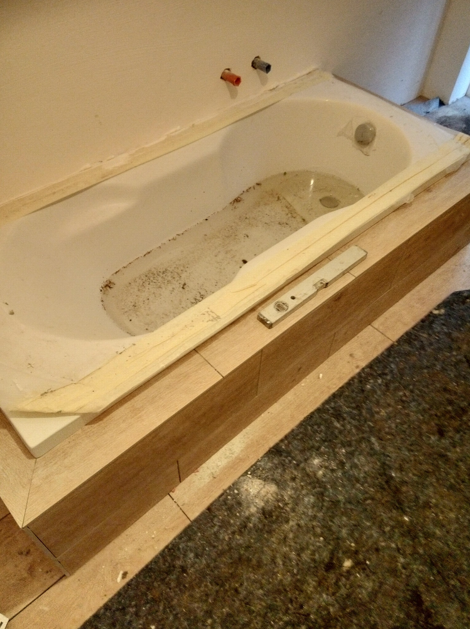 Biała wanna w trakcie obudowy z drewnianych desek, poziomica leży obok. Widoczne przyłącza wodne na ścianie. Etap prac wykończeniowych w łazience.