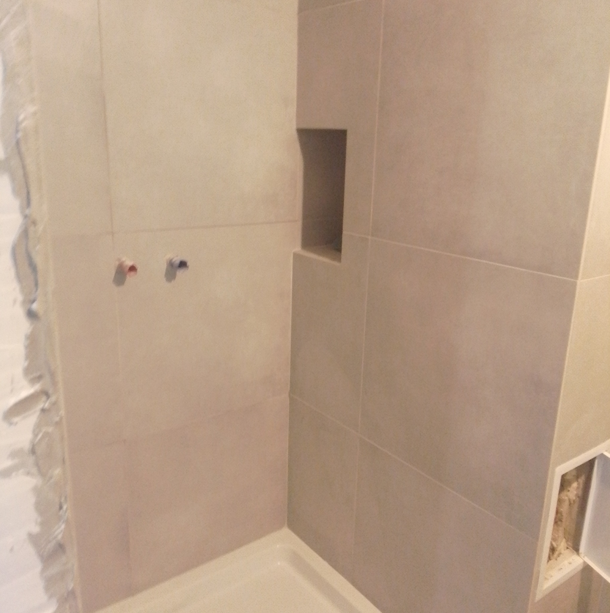 Wykończona wnęka w kabinie prysznicowej z beżowymi, dużymi płytkami. Widoczne wystające rury. Fragment ściany z niedokończonym wykończeniem.