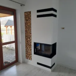 Nowoczesny biały narożny wkład kominkowy z czarnymi akcentami, obok dekoracyjna ściana z drewna, widok z boku na jasne panele podłogowe i drzwi balkonowe.