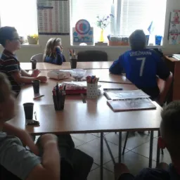 Grupa uczniów siedzi przy stole podczas lekcji w klasie, na ścianie wiszą materiały edukacyjne i zdjęcia.