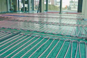 Instalacja ogrzewania podłogowego: rury grzewcze ułożone równolegle na zielonej folii izolacyjnej, przygotowane do zalania wylewką. Widoczne okna i elementy konstrukcyjne budynku w tle.