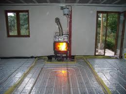 Instalacja ogrzewania podłogowego z widocznym piecem kominkowym typu koza z płomieniami wewnątrz. Rury ogrzewania podłogowego ułożone na folii izolacyjnej, przygotowane do zalania wylewką.