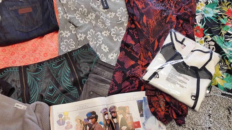 Stos ubrań z różnych materiałów i wzorów, w tym dżinsy Wrangler, bluzka River Island z metką, sukienka ze wzorem skóry węża, przewodnik po modzie na otwartej książce i zapakowana bluzka z czarnymi...