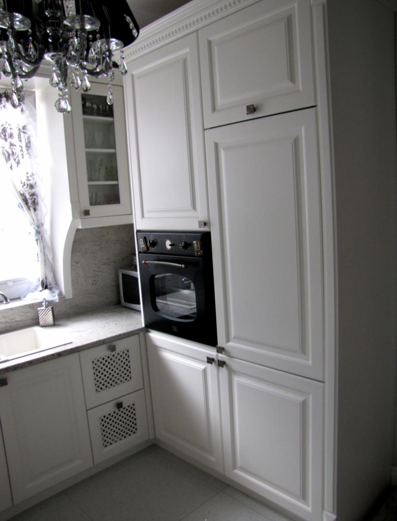 Biała zabudowa kuchenna z piekarnikiem w stylu retro, ozdobnymi szufladami i kryształowym żyrandolem