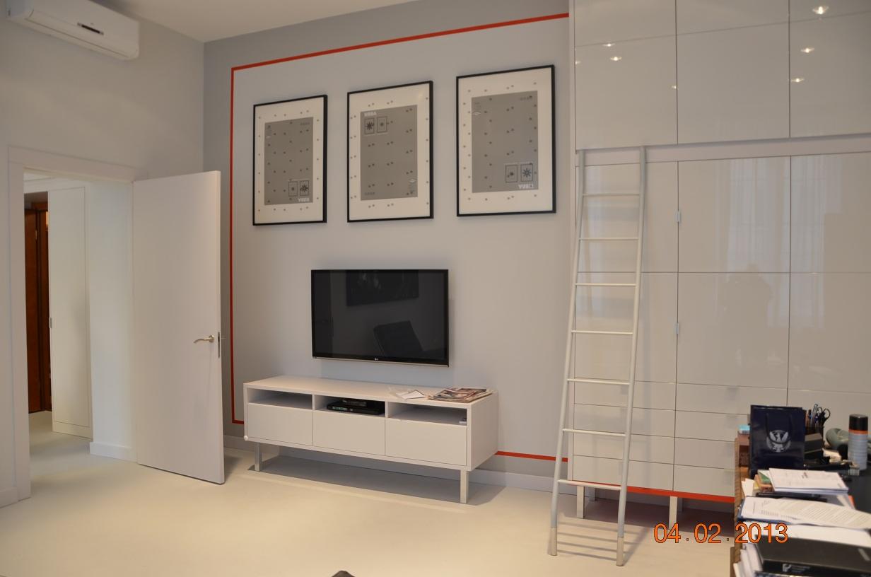 Biały, minimalistyczny salon z szafą na całej ścianie, do której dostawiona jest biała drabina. Na ścianie wiszą trzy obrazy w czarnych ramkach i telewizor nad białą szafką RTV.