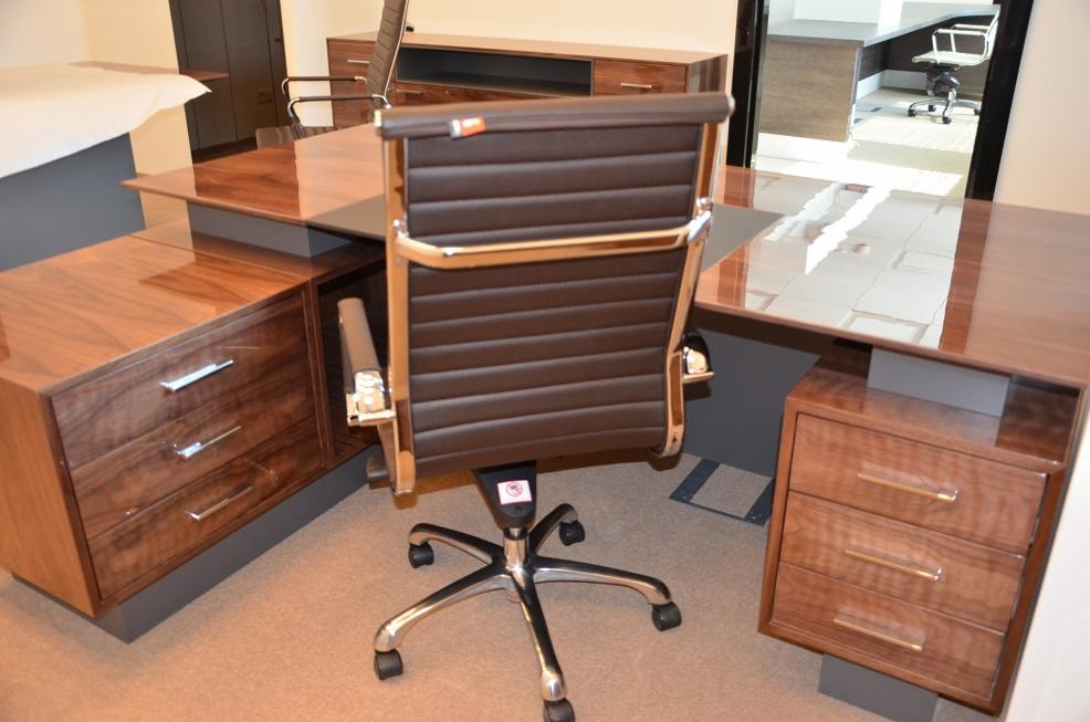 Nowoczesne biurko narożne z drewna orzechowego z szufladami i chromowanym krzesłem biurowym z brązowym skórzanym oparciem, widok z tyłu krzesła, w tle jasne pomieszczenie biurowe.