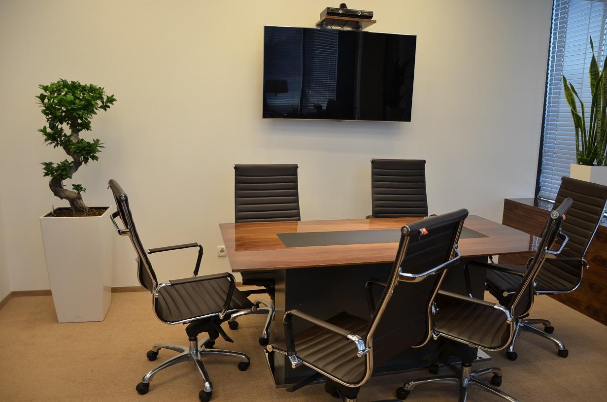 Sala konferencyjna z dużym drewnianym stołem z czarnym wkładem, otoczonym czarnymi krzesłami na kółkach, na ścianie telewizor i roślina w doniczce.