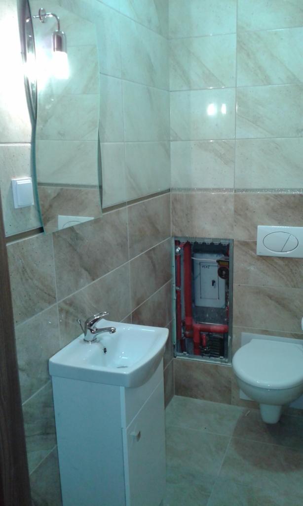 Wykończona łazienka z beżowymi płytkami imitującymi marmur, białą szafką pod umywalką i toaletą. Widoczne czerwone rury instalacyjne za otwartym panelem rewizyjnym.