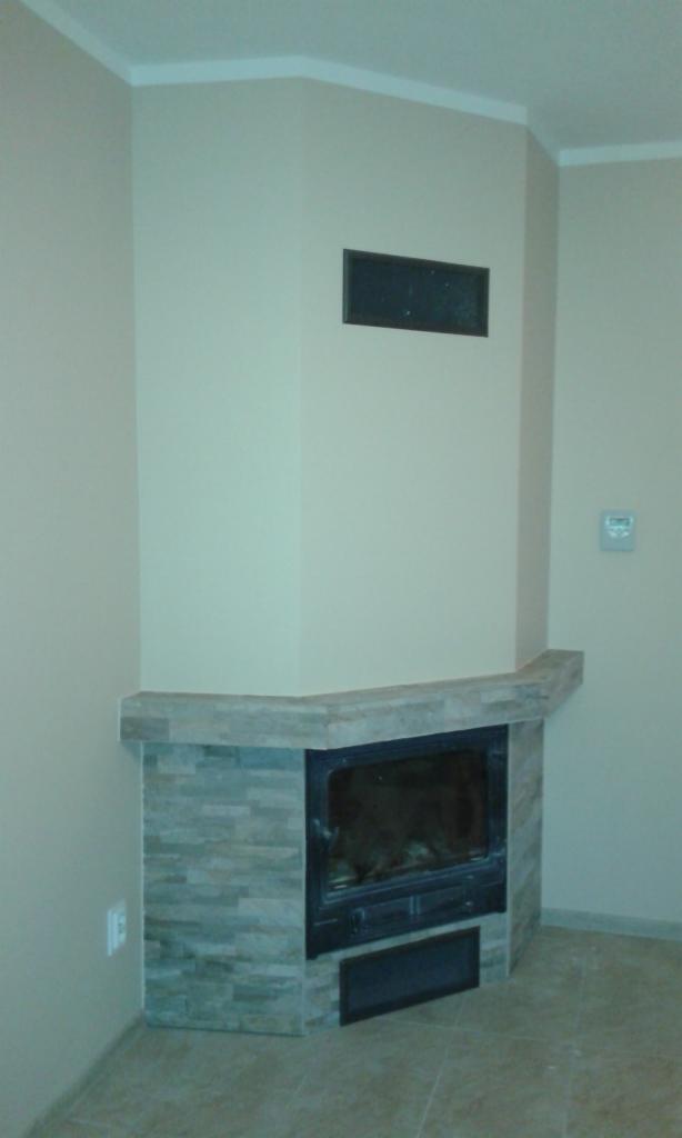 Kątowy kominek obudowany płytkami imitującymi kamień, z czarnym wkładem kominkowym i kratką wentylacyjną powyżej, w jasnym wnętrzu.