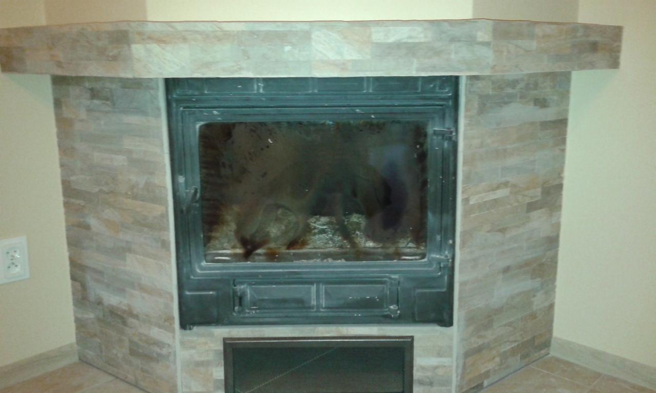 Obudowa kominka wykończona płytkami imitującymi kamień, z czarnym wkładem kominkowym o zabrudzonej szybie.