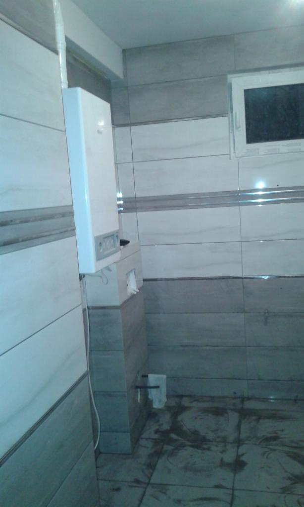 Biały piec gazowy zamontowany na ścianie wyłożonej szaro-białymi płytkami w łazience z widocznym fragmentem okna i niedokończoną podłogą.