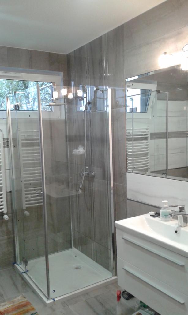 Nowoczesna kabina prysznicowa ze szklanymi ściankami i chromowanymi okuciami, zamontowana w łazience wykończonej szarymi płytkami imitującymi kamień, z widoczną umywalką z białymi szafkami i lustrem.