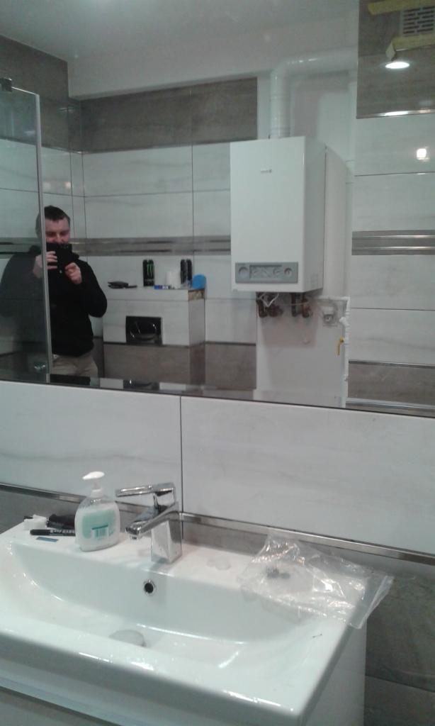 Biały piec gazowy zamontowany na ścianie w łazience z białymi i szarymi płytkami, widoczne rury i instalacja, umywalka z baterią i dozownikiem mydła, odbicie fotografa w lustrze.