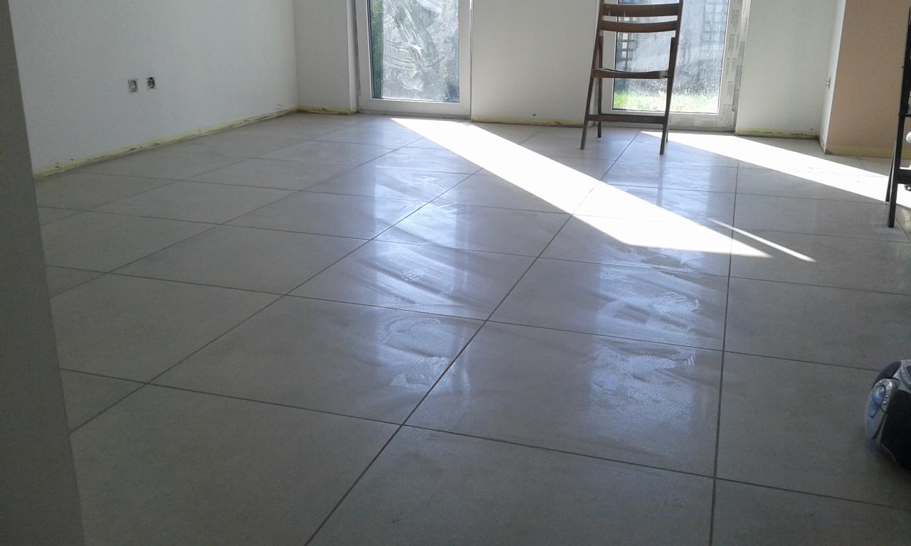 Świeżo położona, jasnoszara podłoga z płytek ceramicznych w pustym pokoju, z widoczną smugą światła słonecznego i krzesłem w tle, taśma malarska zabezpiecza ściany.
