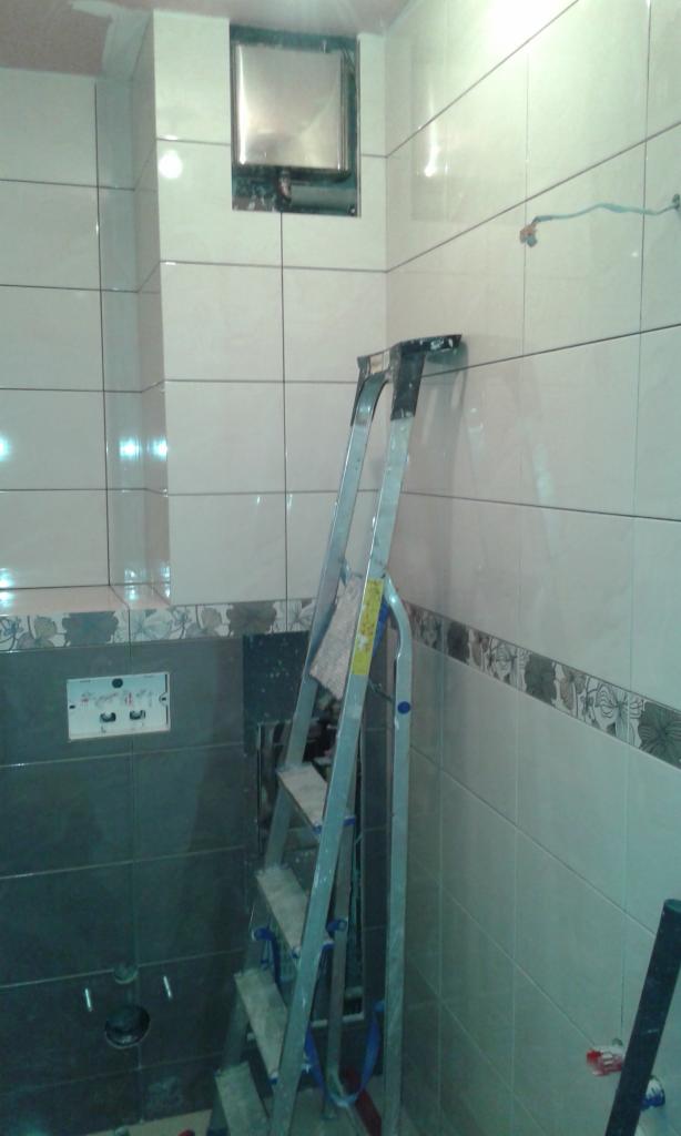 Łazienka w trakcie układania płytek; widoczna drabina oparta o ścianę z częściowo ułożonymi białymi płytkami i dekoracyjnym pasem, a także szare płytki w dolnej części ściany z przygotowanymi...