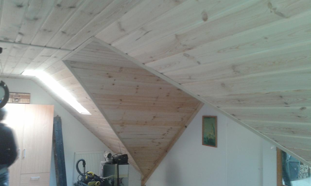 Drewniana boazeria pokrywająca skośny sufit na poddaszu, z widocznym oknem dachowym i zielonym obrazkiem na białej ścianie w tle.