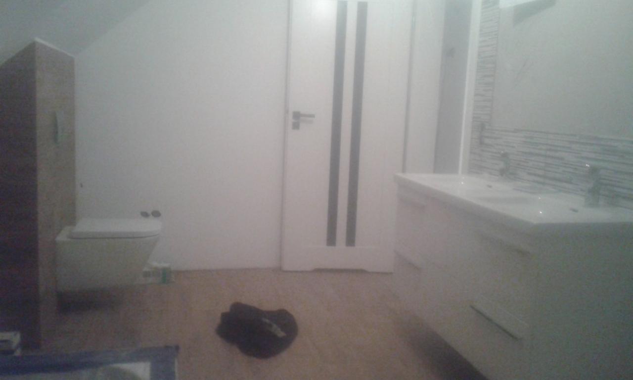 Nowoczesna łazienka w trakcie wykańczania: podwieszana toaleta, biała szafka z umywalkami i armaturą, białe drzwi z ciemnymi paskami, brązowe panele podłogowe, widoczne ślady po remoncie.