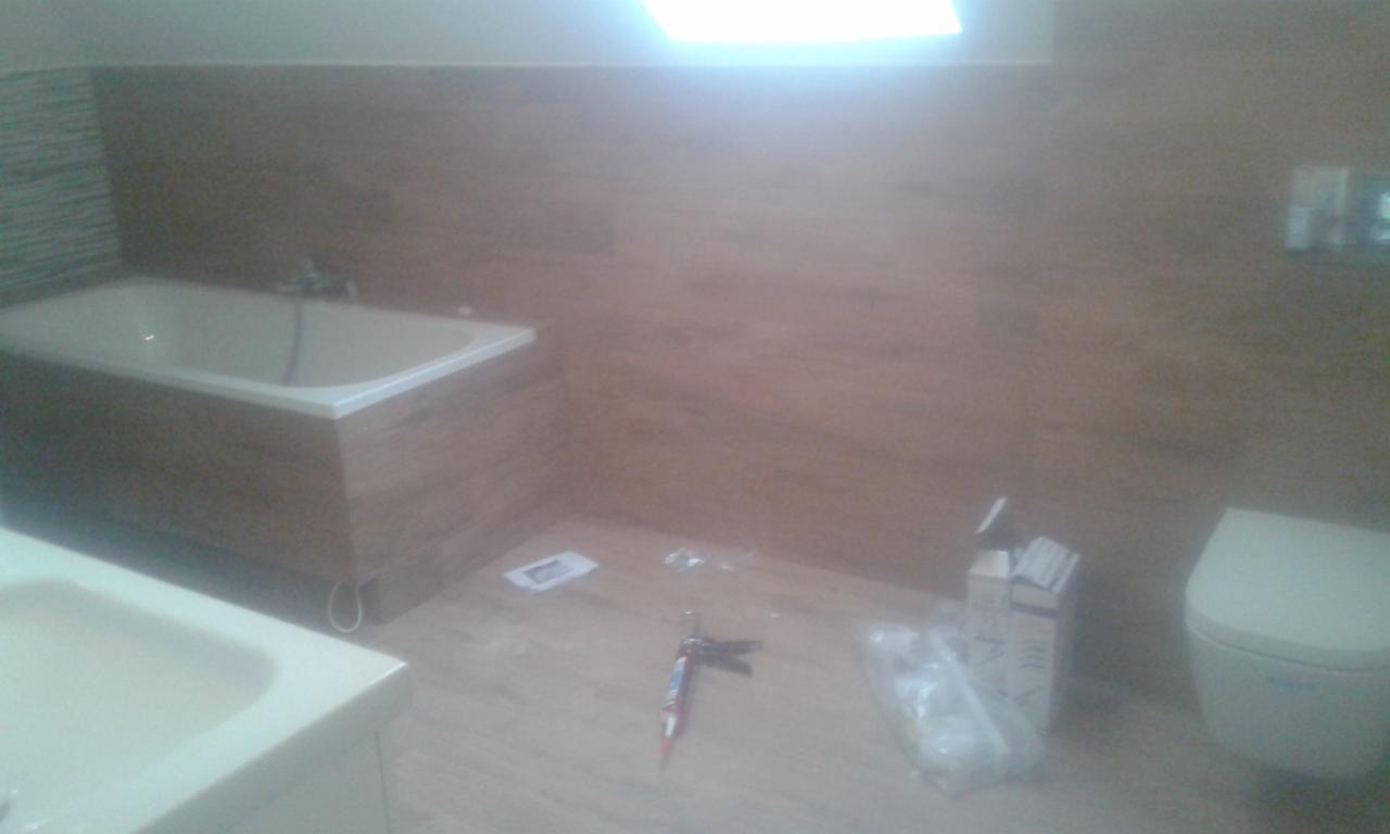 Montaż płytek imitujących drewno w łazience, widoczna wanna z obudową, umywalka, toaleta oraz narzędzia i materiały budowlane na podłodze.