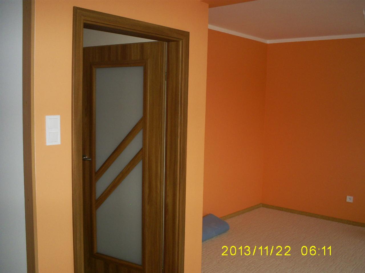 Pomalowane na pomarańczowo ściany pokoju z otwartymi drewnianymi drzwiami z szybą o geometrycznym wzorze, widoczny fragment podłogi i niebieska poduszka.