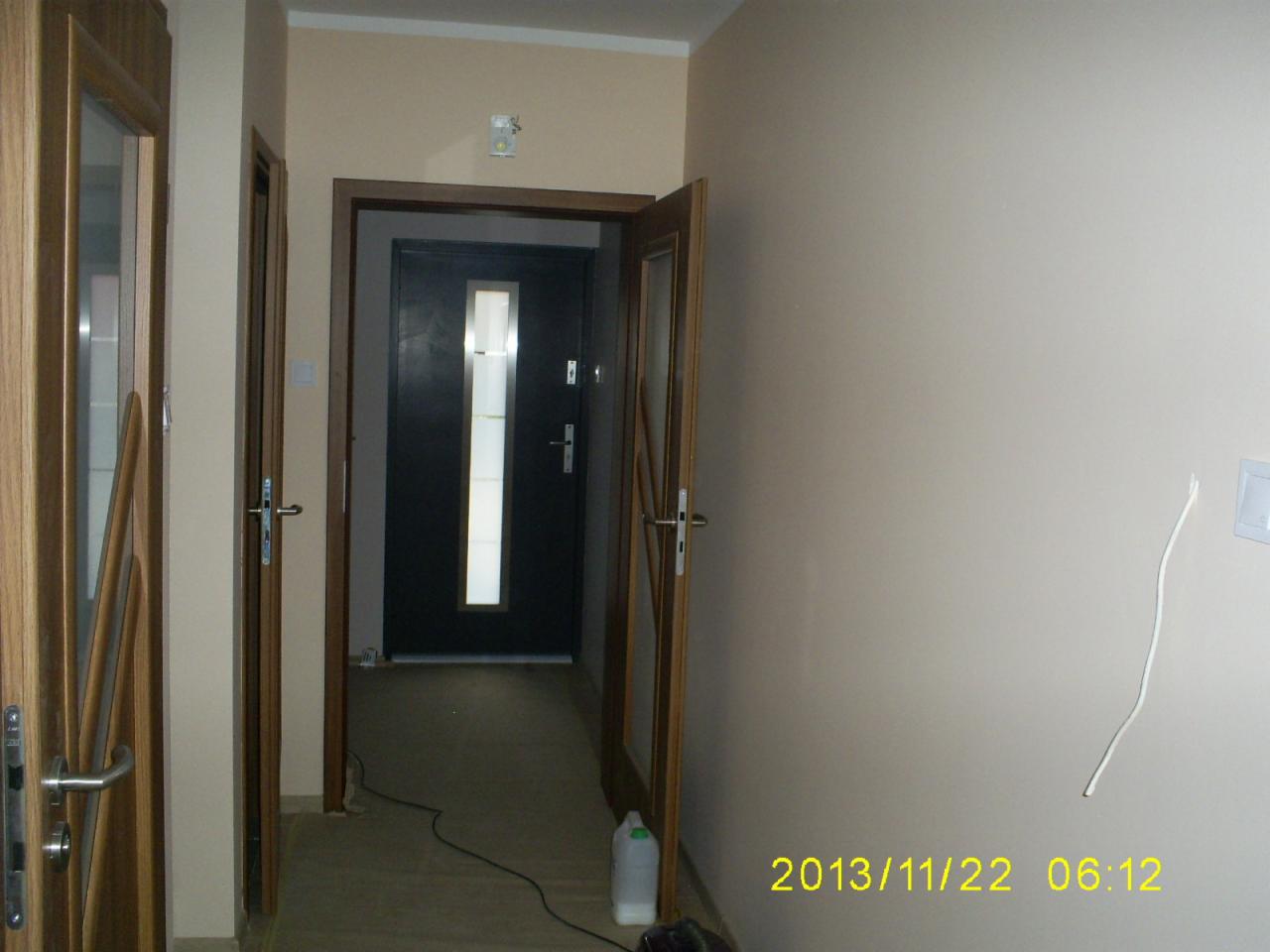 Korytarz w trakcie prac wykończeniowych: widoczne drzwi wewnętrzne z drewnianą ramą i szklanym panelem, otwarte drzwi wejściowe z czarną ramą i podłużnym oknem, ściany w odcieniach beżu, kabel...