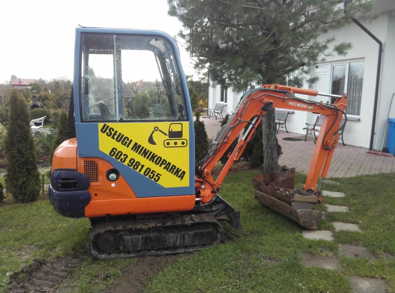 Pomarańczowo-niebieska mini koparka Hitachi z żółtym napisem 'Usługi Minikoparka 603 981 055' na tle zielonego trawnika i białego domu.