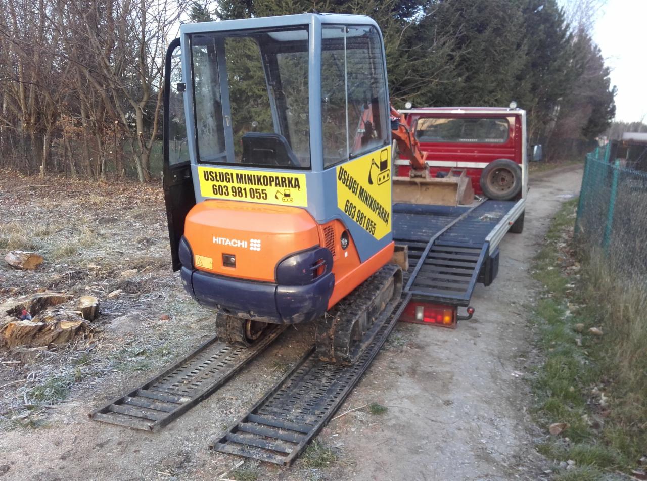 Pomarańczowo-niebieska mini koparka Hitachi na rampach wjazdowych, gotowa do transportu na platformie samochodu ciężarowego. Na koparce naklejka z numerem telefonu.